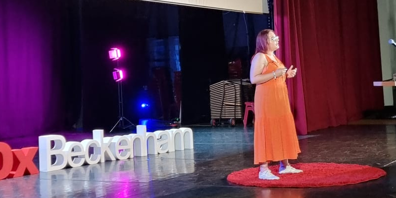 Sonay on stage at her TedX talk in Beckenham. Behind her on a screen is the word 20000 indicating how many more negative comments a neurodiverse child gets by age 12 compared to their neurotypical peers.